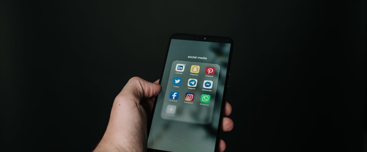 Close-up of a hand holding a smartphone displaying various social media app icons on a dark background.