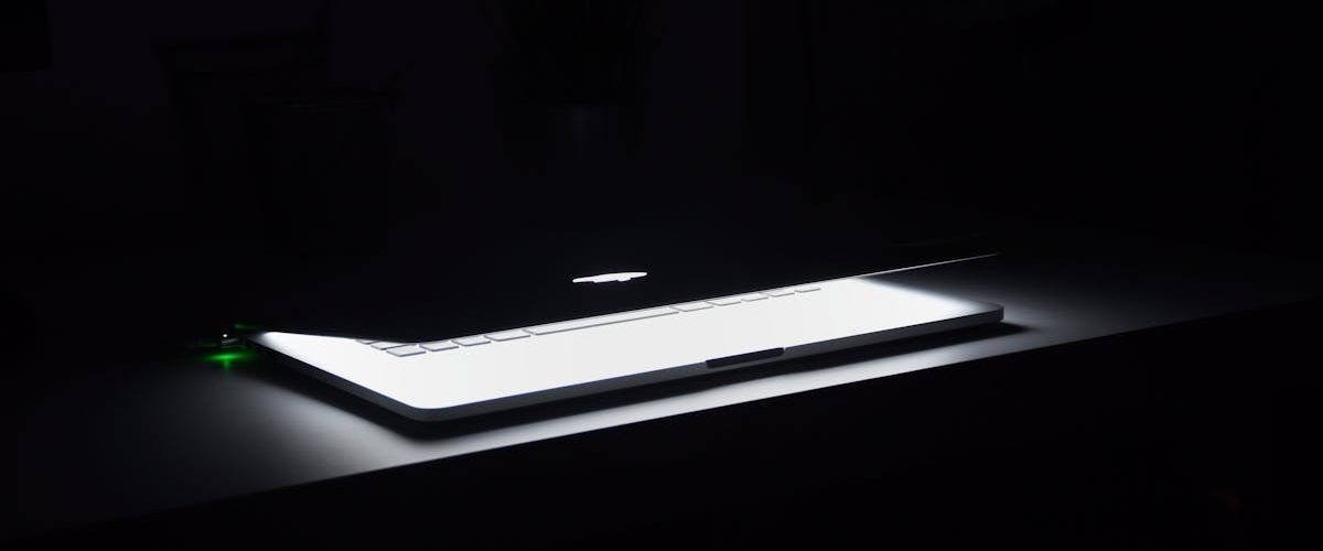 A dramatic, low-light image of a slightly open laptop with glowing keys on a dark desk.
